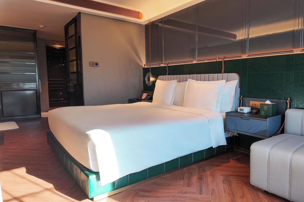 A modern hotel room with a large bed, a bedside table with a lamp, a small seating area, and a view of the city through the window.