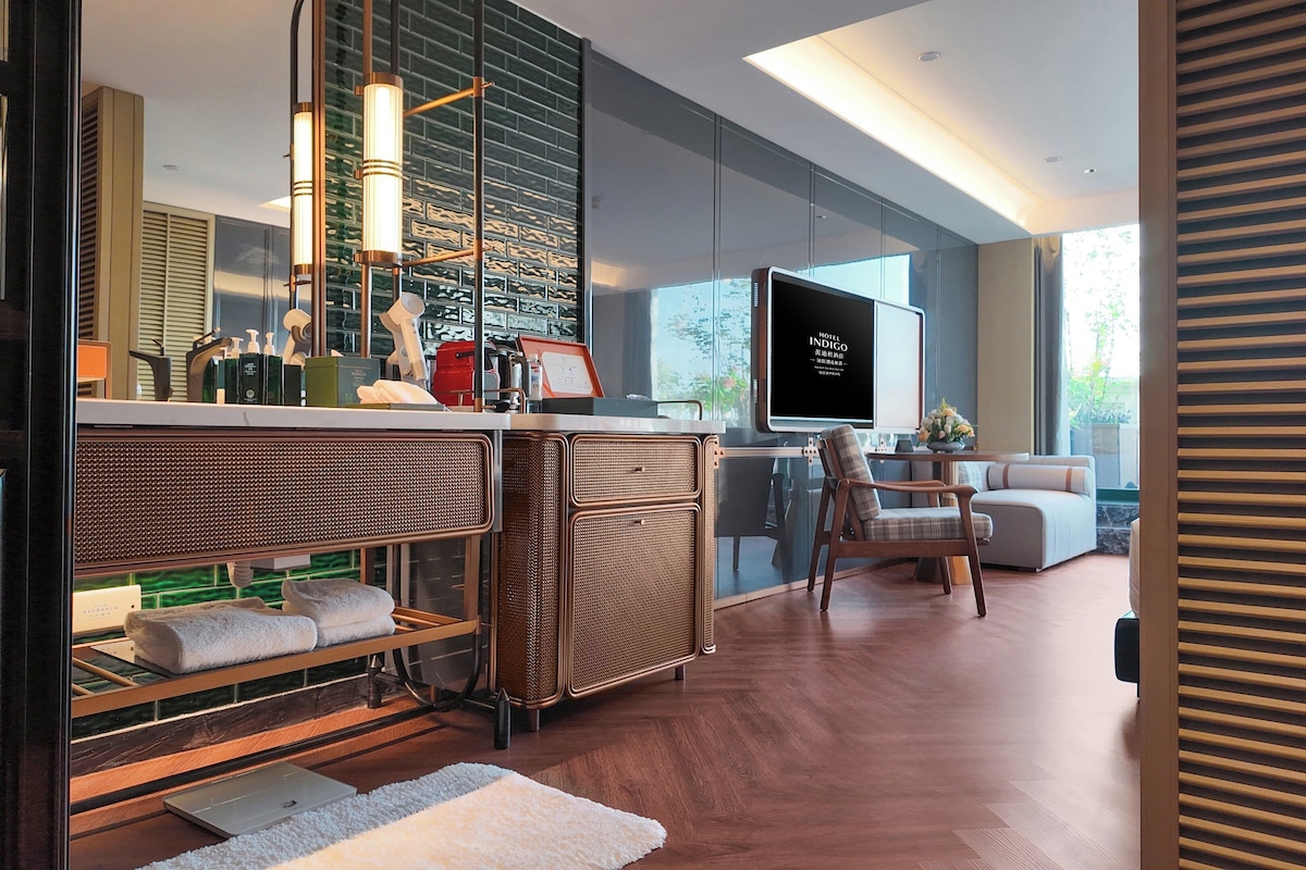 Una habitación de hotel moderna con un televisor grande, una zona de estar con sofá y sillón, un espacio tipo bar con botellas y una computadora portátil, y un ventanal con vistas.