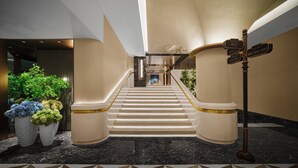 Una imponente escalera con peldaños de mármol y barandales dorados, que conduce a un vestíbulo de diseño moderno, con un arreglo de plantas y señalización.
