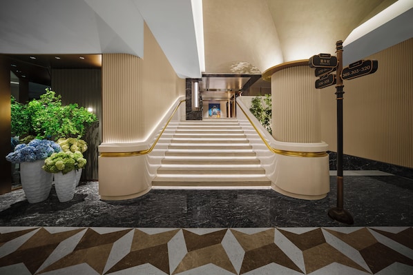 Un magnifique escalier en marbre, avec des marches et des rampes dorées, mène à un hall d’entrée au design moderne, agrémenté d’une exposition de plantes et de panneaux indicateurs.