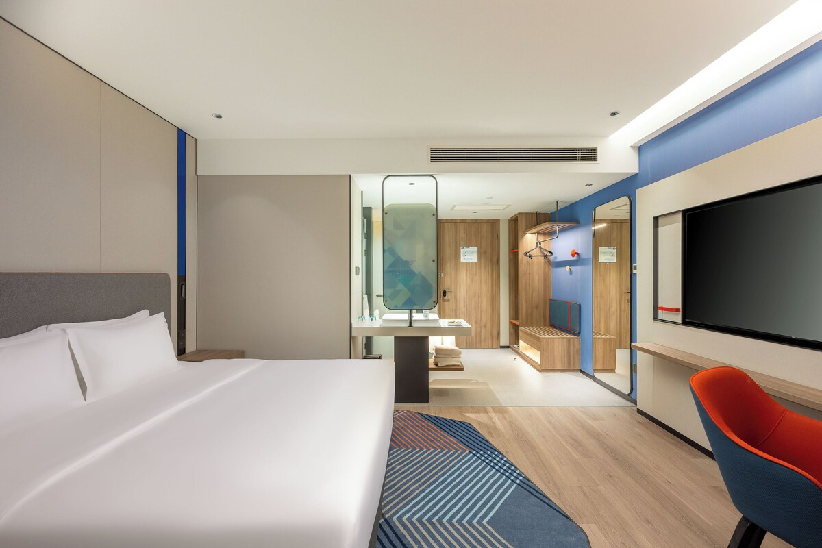 A modern hotel room with a large bed, a TV, a desk, and a bathroom visible through an open door.