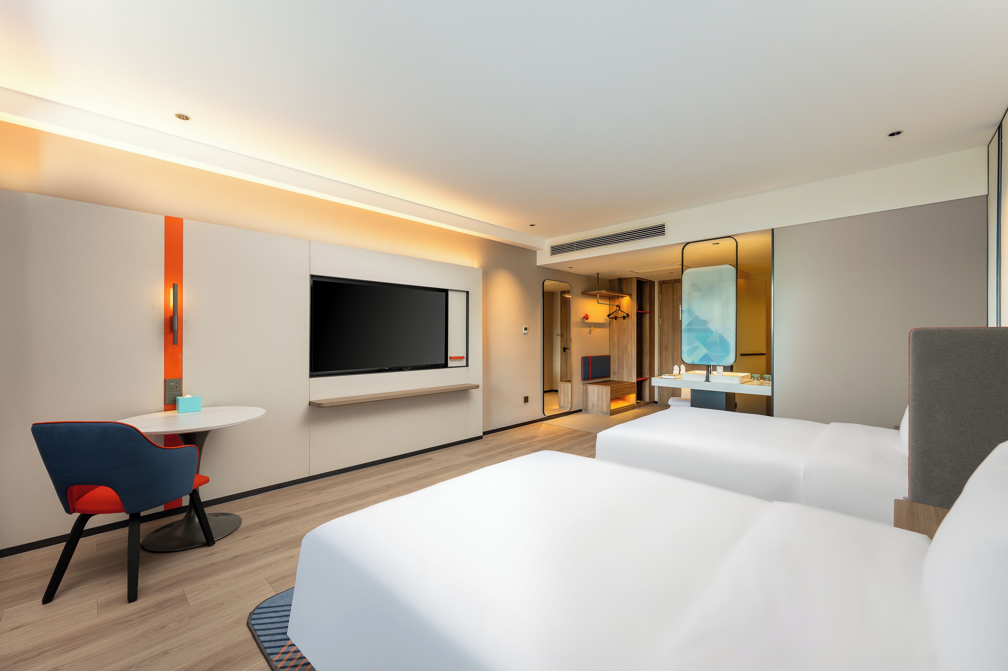 Habitación de hotel con televisor de pantalla plana, una mesita con una silla, dos camas, baño con espejo y un ventanal grande.