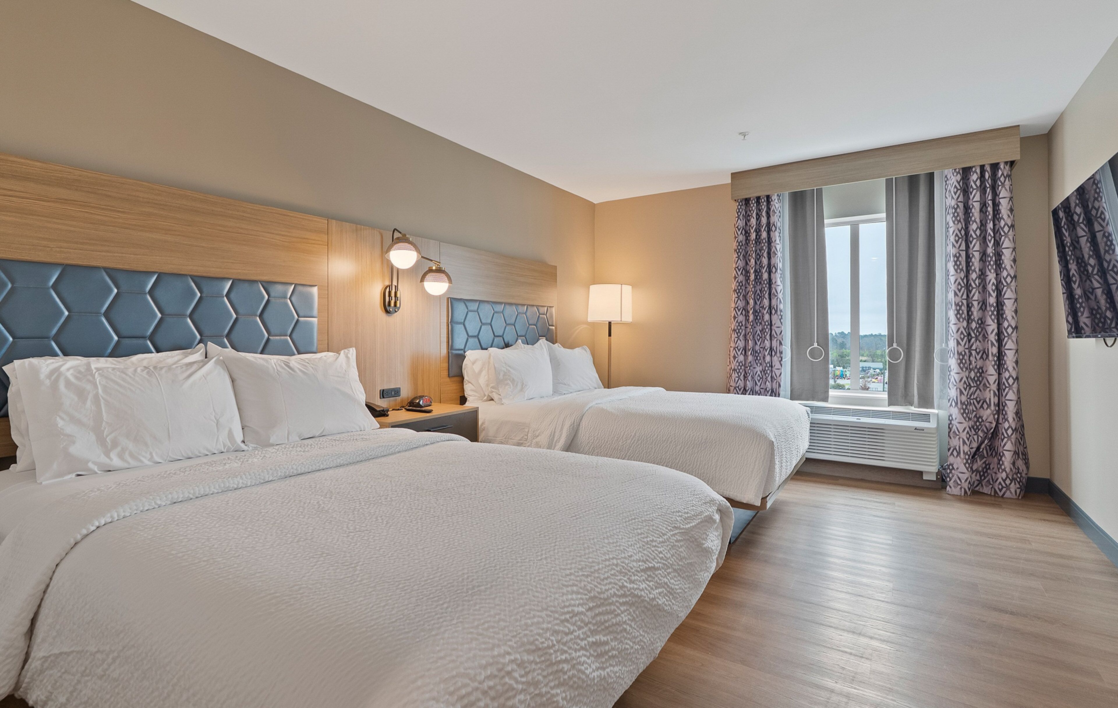 A hotel room with two beds, a wooden headboard, a nightstand with a lamp, and a large window with curtains.