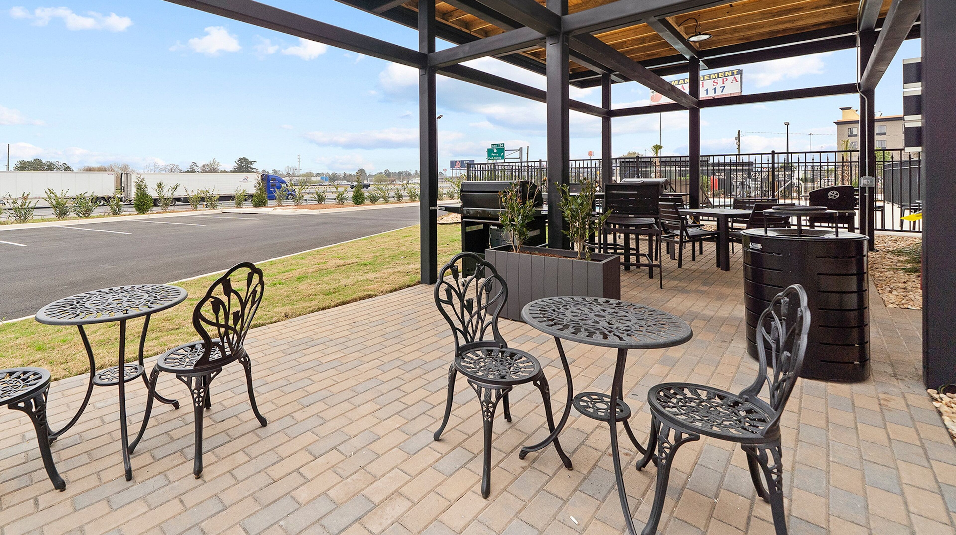 An outdoor patio area with metal tables and chairs, a barbecue grill, and a view of a road and distant buildings.