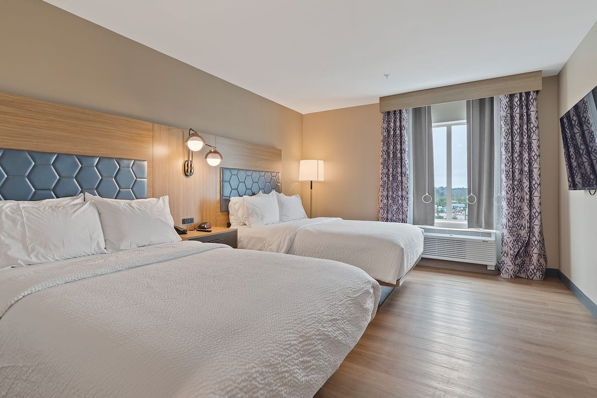 A hotel room with two beds, a wooden headboard, a nightstand with a lamp, and a large window with curtains.