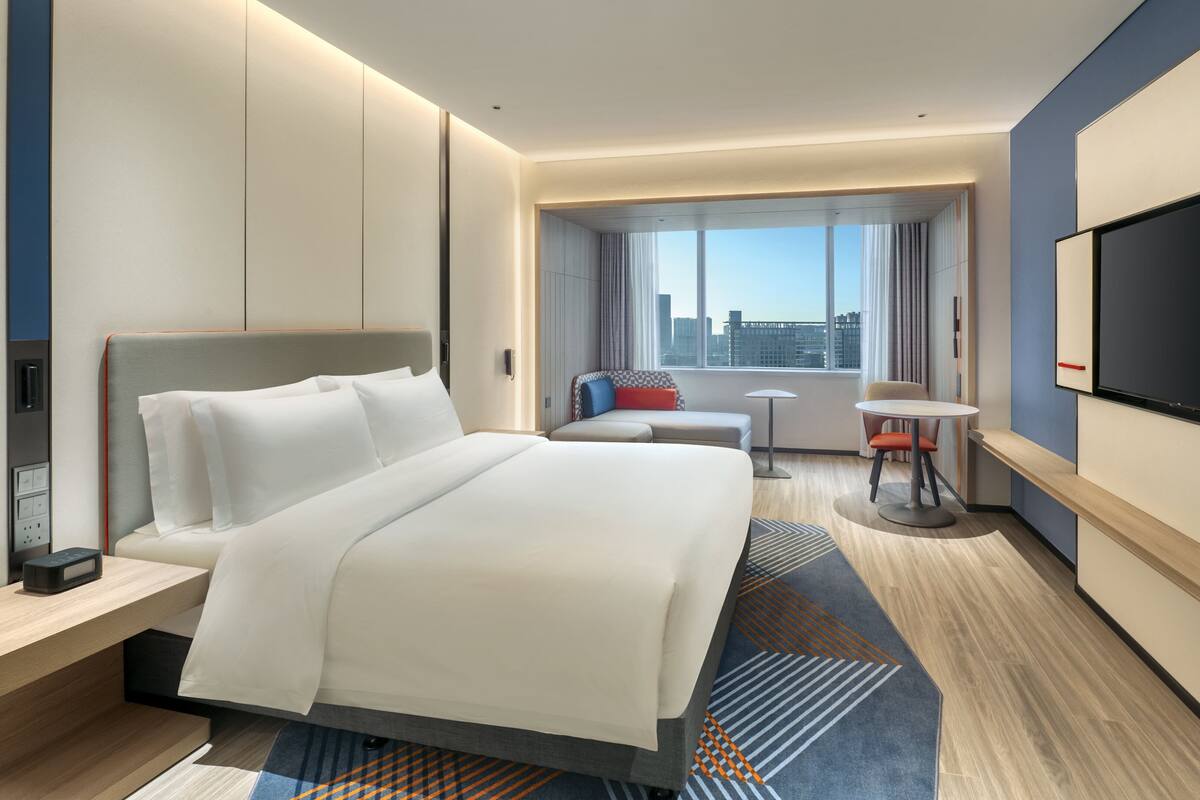 A modern hotel room with a large bed, a desk, a chair, a small table, and a TV.