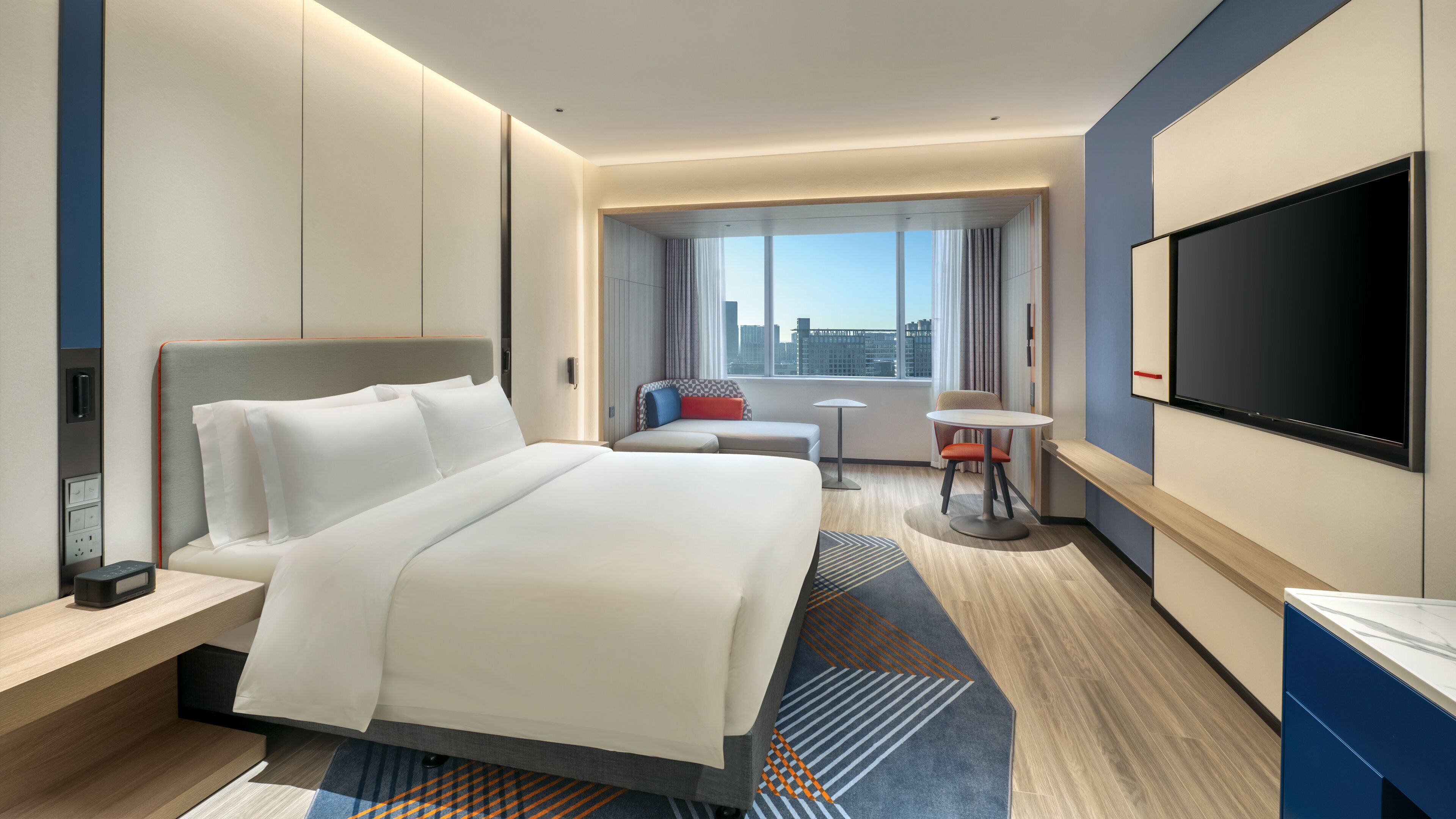 A modern hotel room with a large bed, a desk, a chair, a small table, and a TV.