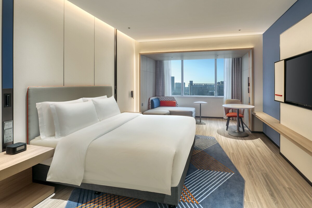 A modern hotel room with a large bed, a desk, a chair, a small table, and a TV.