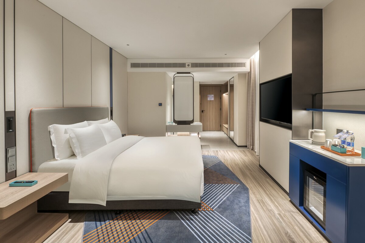 A modern hotel room with a large bed, bedside table, TV, and a small desk area with amenities.
