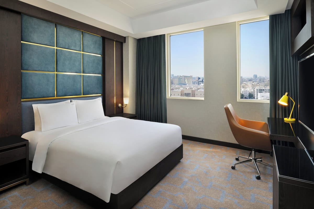 A hotel room with a large bed, a desk with a chair, a window with a city view, and a large wall panel.