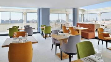 Un comedor moderno con varias mesas y sillas, ventanas amplias que ofrecen vista a la ciudad, y sillas tapizadas en tonos verde y naranja.