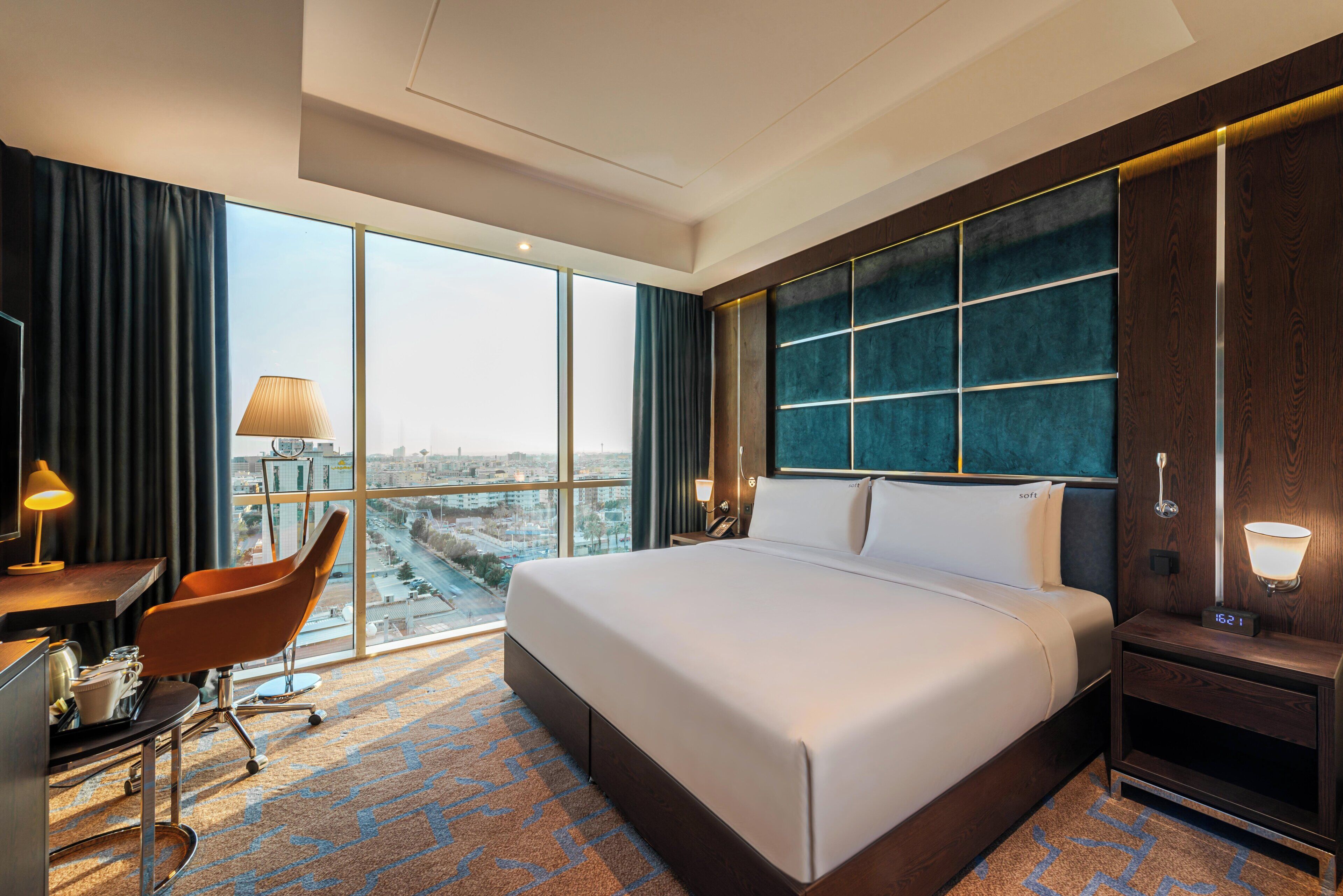 A hotel room with a large bed, a desk with a chair, a nightstand, and a view of the city through large windows.