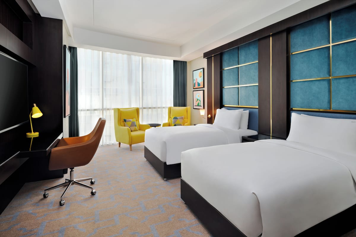A hotel room with two beds, a desk, a chair, and a large window.