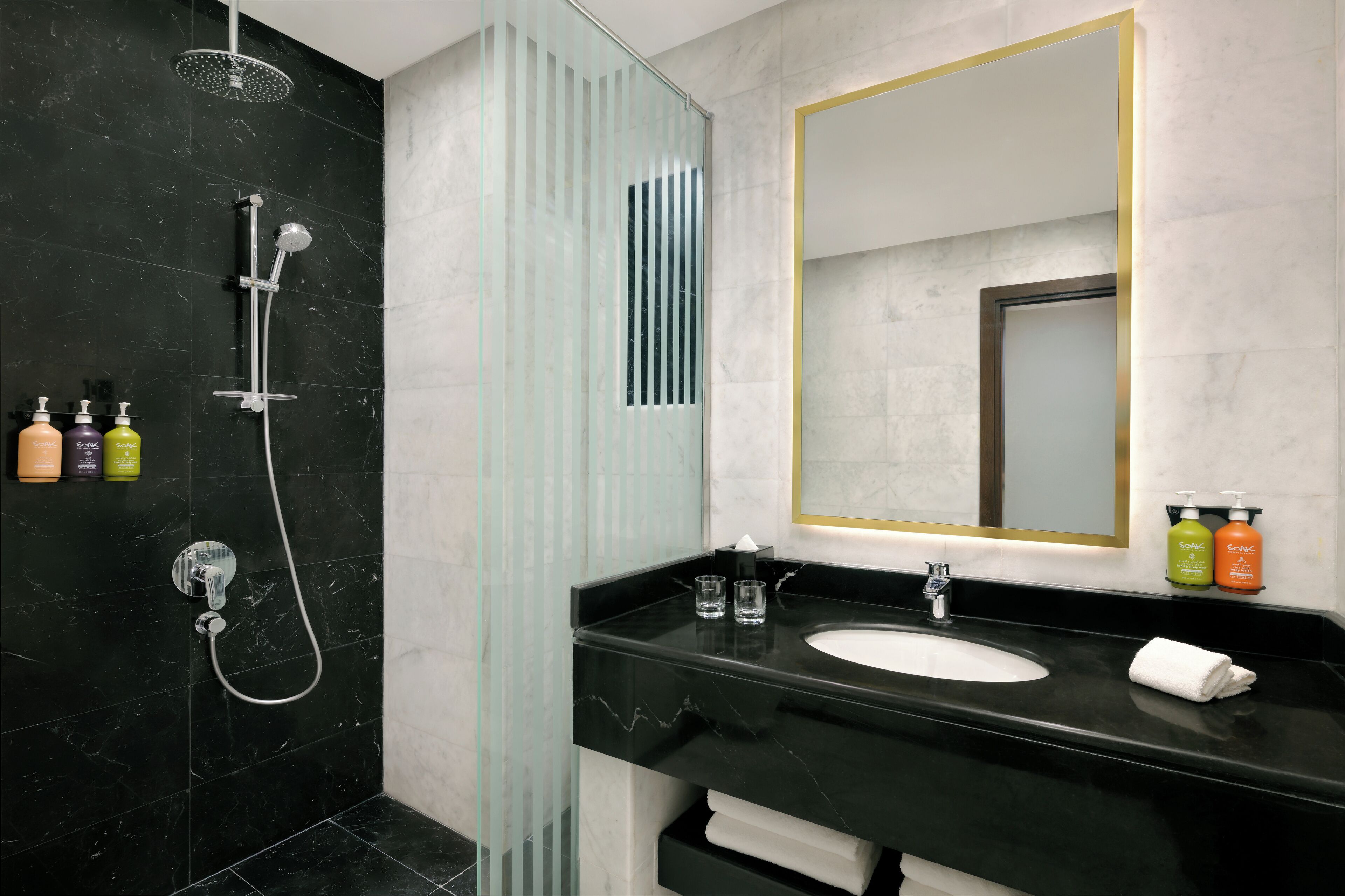 Un baño moderno con ducha de mármol negro, mampara de cristal, espejo grande con marco dorado y encimera negra con lavabo y artículos de aseo.