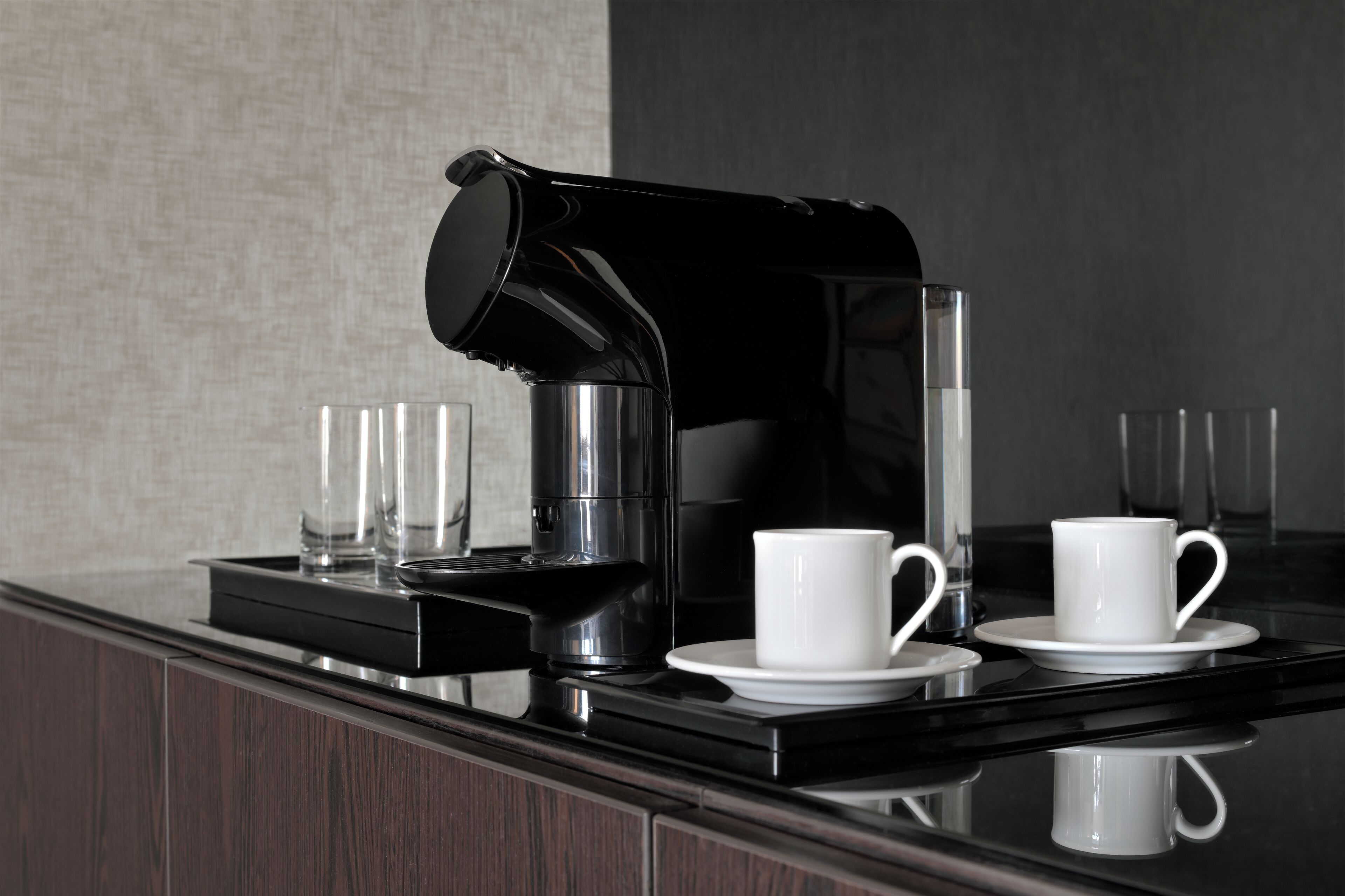 A modern coffee station with a black coffee maker, two white cups, and two clear glasses on a reflective black countertop.