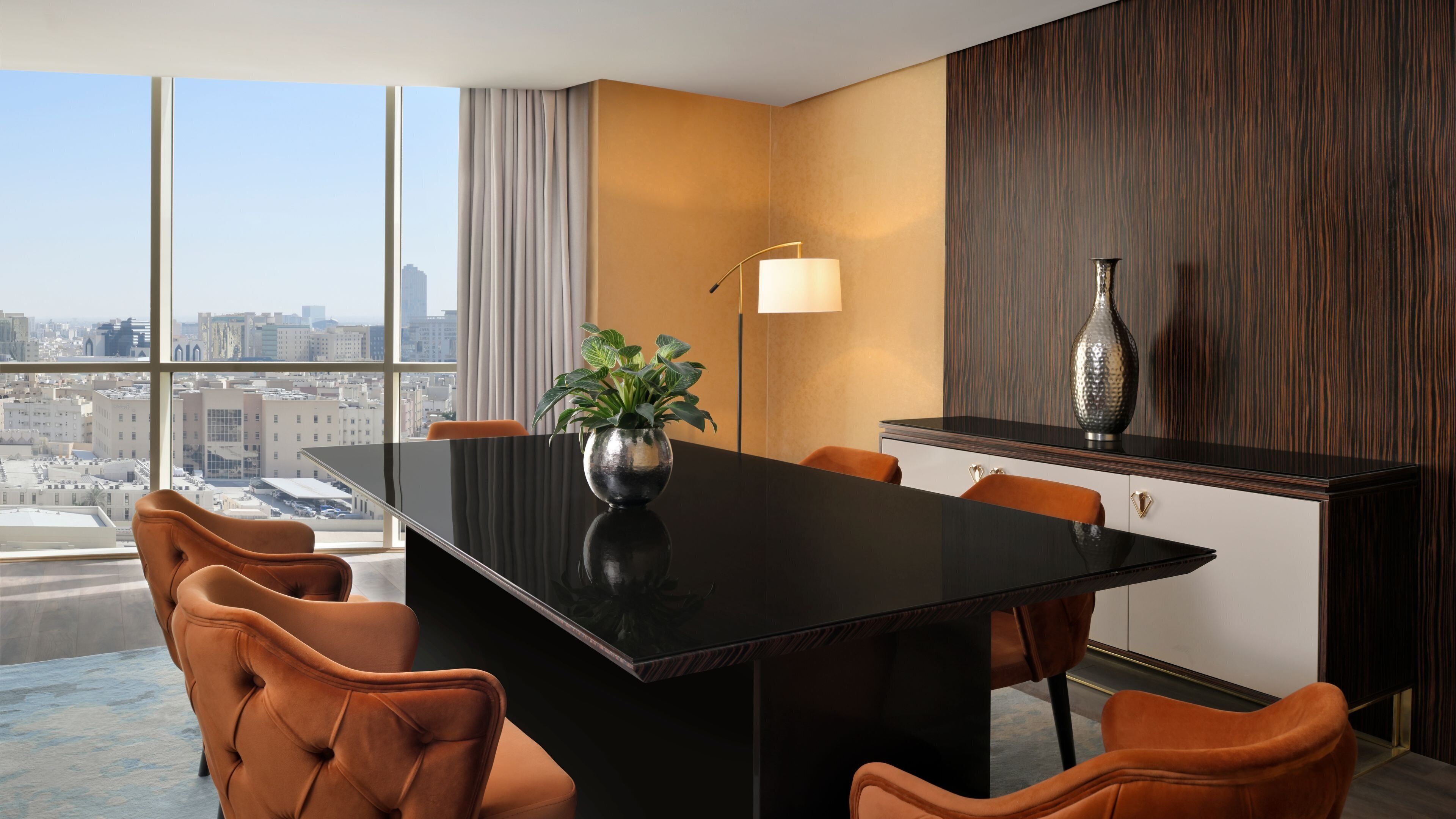 A modern hotel room with a large dining table, orange chairs, a potted plant, a lamp, and a decorative vase.