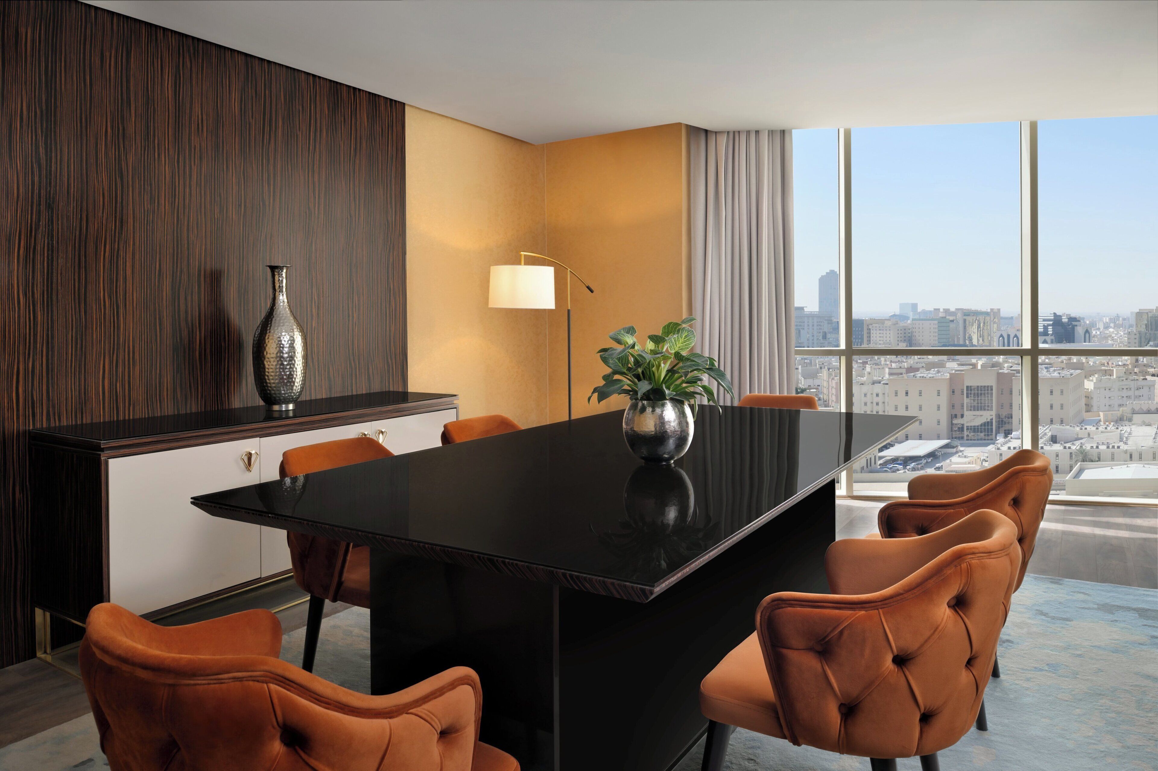 Un coin repas moderne avec une table noire, des chaises orange, un vase avec une plante et une lampe.