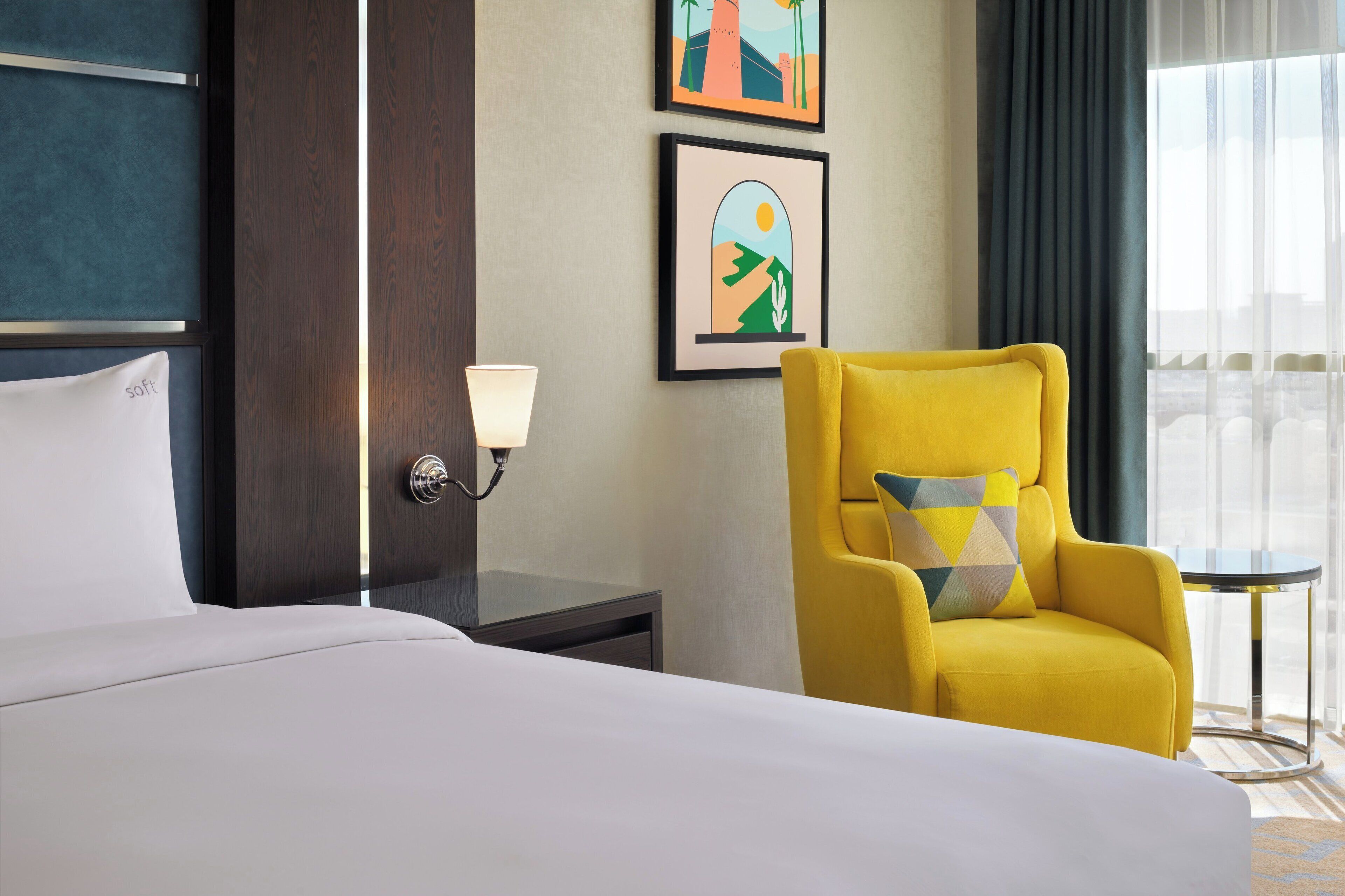Une chambre d’hôtel avec un lit, un fauteuil jaune, une petite table et deux œuvres d’art encadrées accrochées au mur.