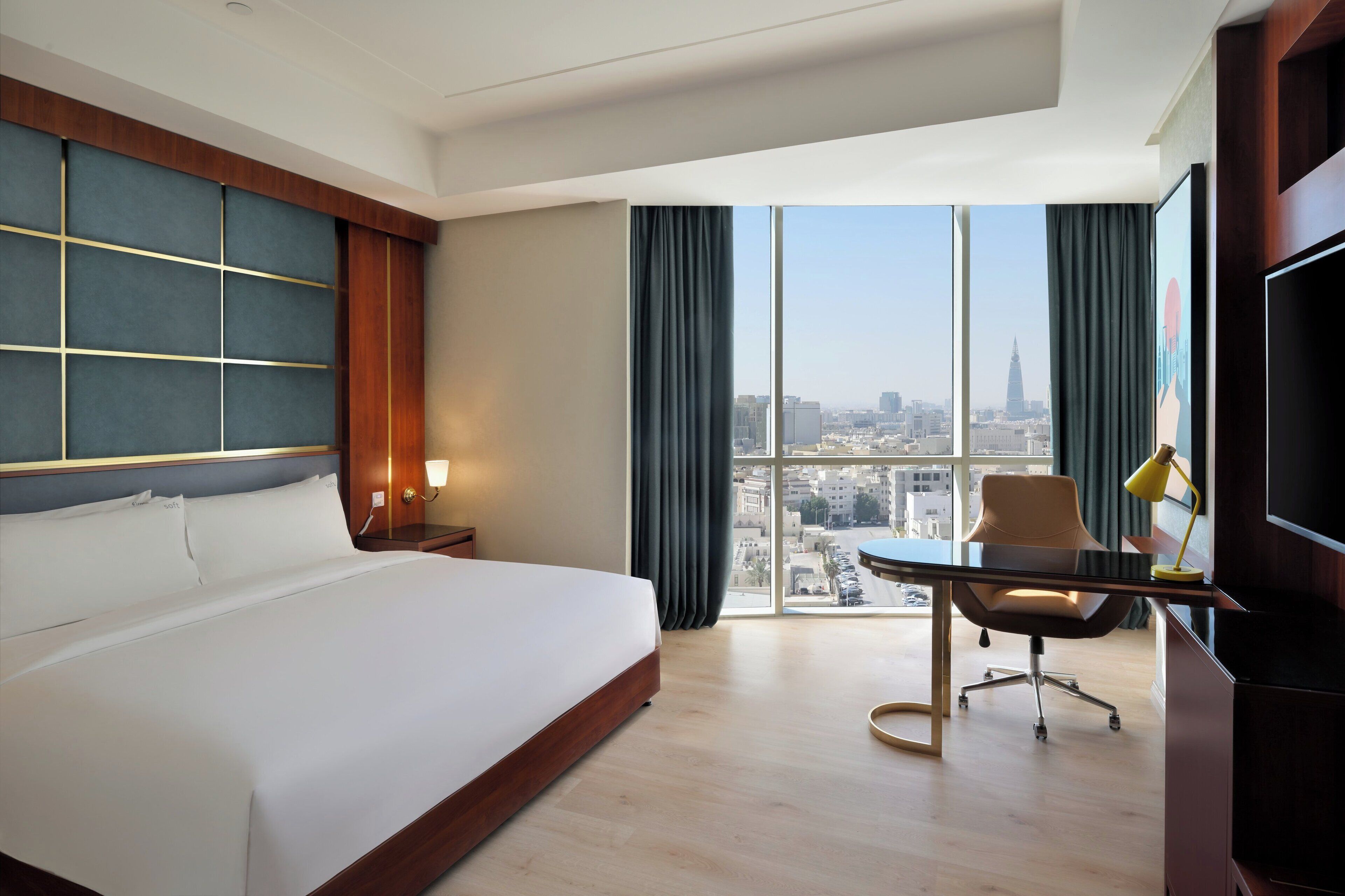 A hotel room with a large bed, a desk with a chair, a TV, and a view of a cityscape through large windows.