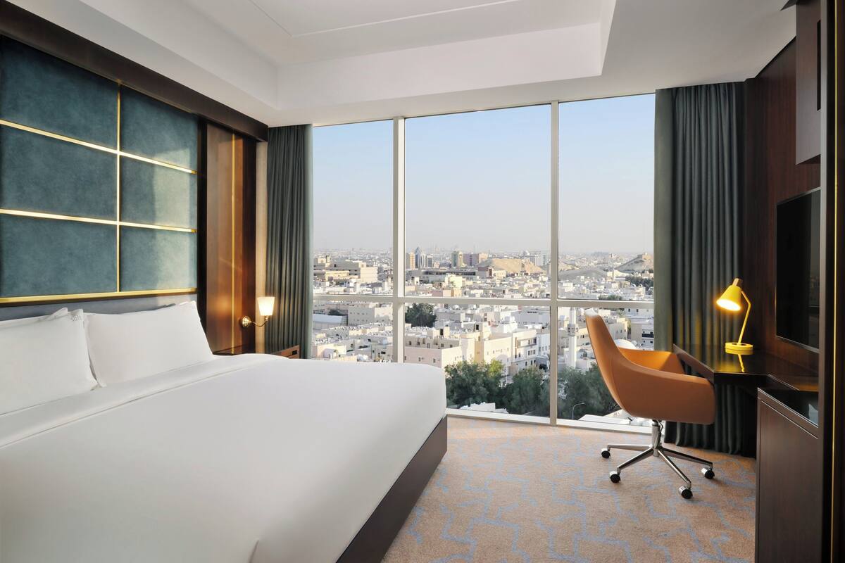 A hotel room with a large bed, a desk with a chair, a TV, and a city view.