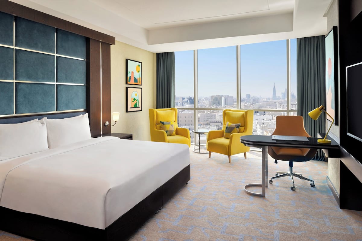 A hotel room with a large bed, two yellow armchairs, a small round table, a desk with a chair, and a large window offering a city view.