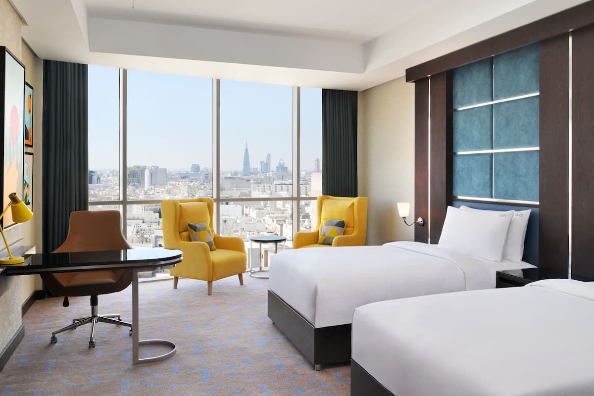 A hotel room with two beds, a desk, a chair, and a view of a cityscape.