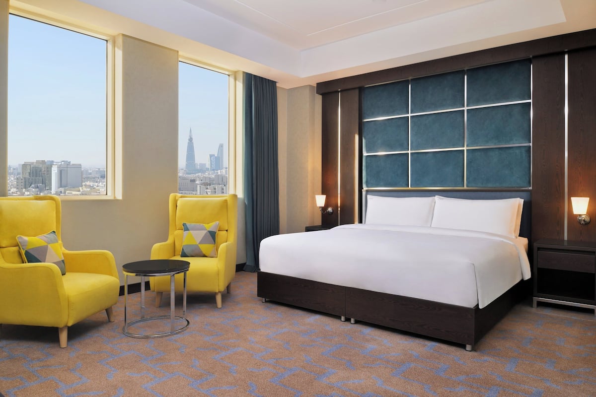A hotel room with a large bed, two yellow armchairs, a small table, and a view of the city skyline through large windows.