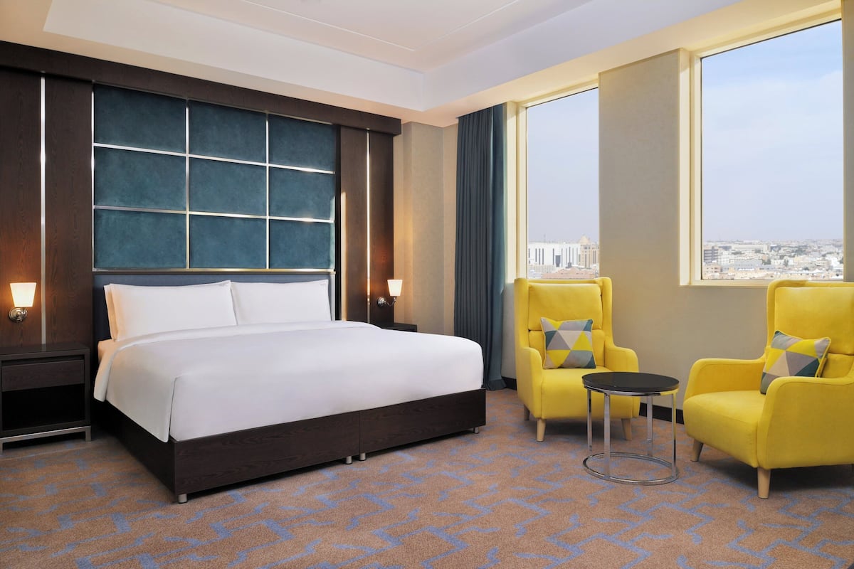 A hotel room with a large bed, two yellow armchairs, a small table, and a view of the city through large windows.