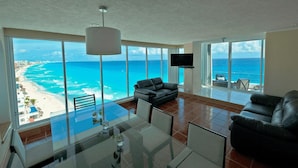 A spacious living room with a large glass table, black leather sofas, and a TV. It features a stunning ocean view through large windows. - Beachfront – 11BR/9Bth – Penthouse Combo #3120 (Cancun)