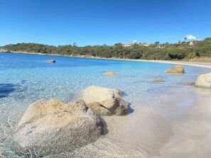 Une plage à l’eau claire, bordée de gros rochers, d’où l’on aperçoit au loin des maisons sur une colline.