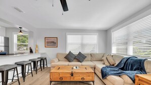 A modern living room with a sofa, coffee table, and bar stools. There is a window with blinds, a framed picture on the wall, and a kitchen area in the background. - Winter Getaway Stuart - Bring your truck, boat or RV - Large Fenced Yard (Stuart)