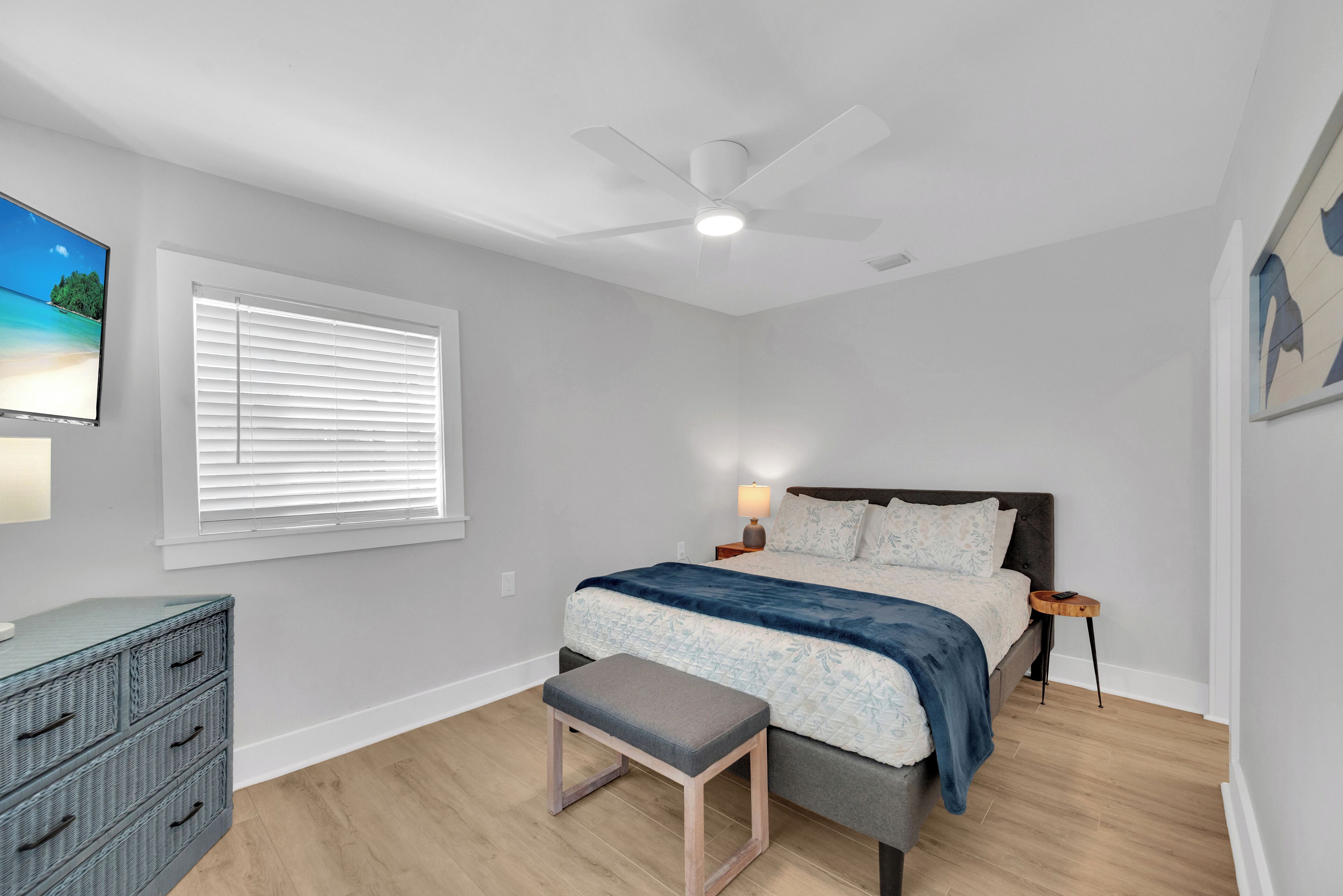 Une chambre à coucher comprenant un lit, une table de chevet, une commode, un banc, un ventilateur de plafond et un téléviseur fixé au mur.