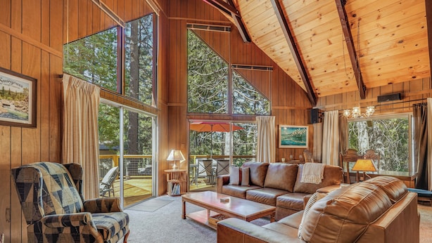 A spacious living room with wooden walls and large windows, a sofa, a chair, and a coffee table. - Arnold Chalet Near Town & Calaveras Big Trees! (Arnold)