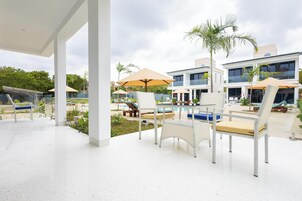 Un área de piscina moderna al aire libre con sillones de descanso y sombrillas, rodeada de palmeras y un cielo despejado.