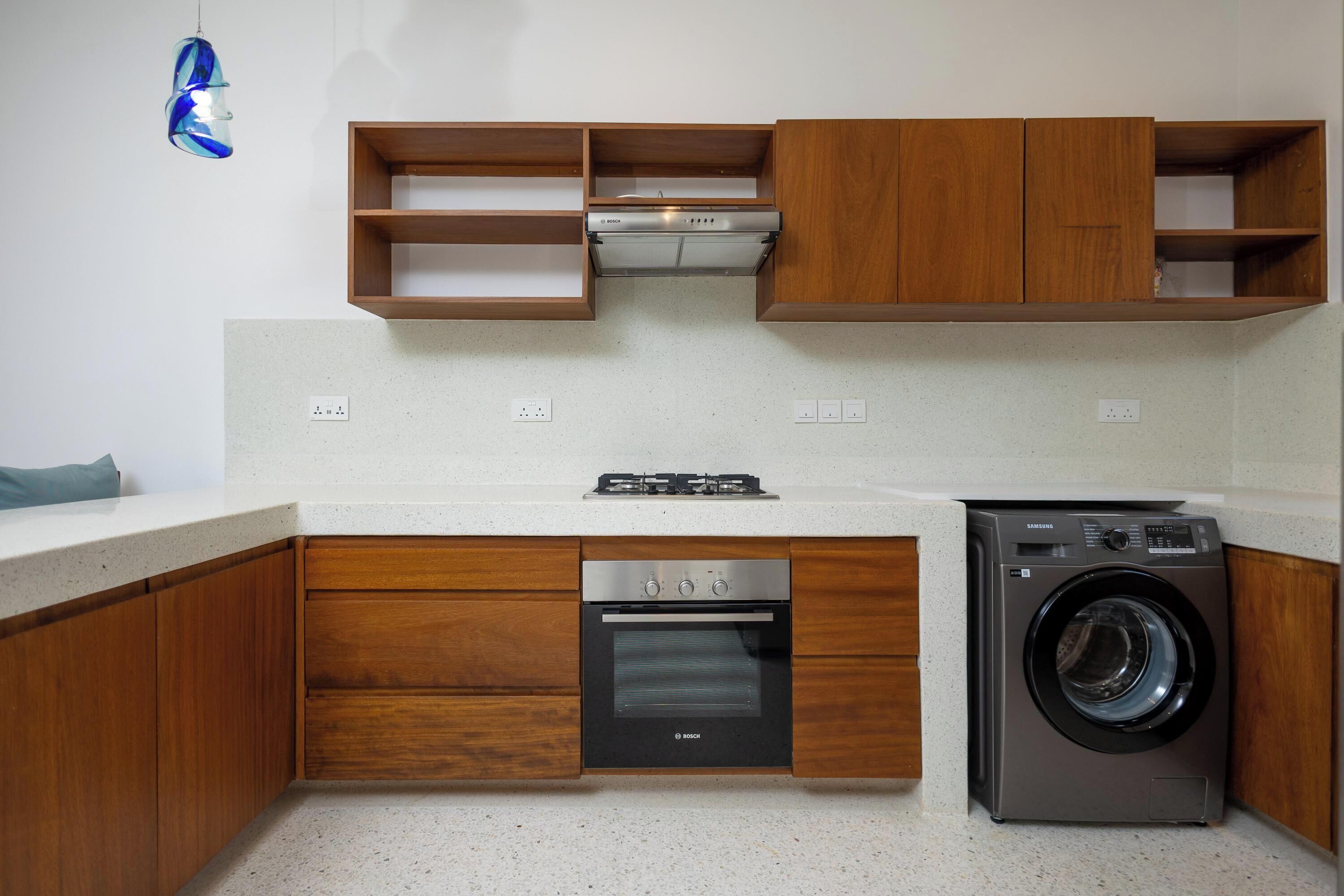 A compact kitchen with wooden cabinets, a Bosch oven, a washing machine, and a blue hanging light fixture.
