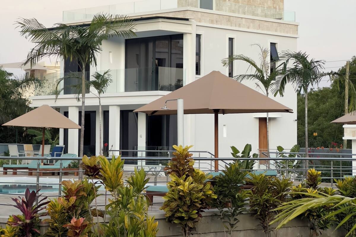 A modern building with a flat roof, large windows, and a balcony. There are palm trees and outdoor seating with umbrellas.