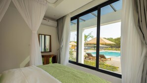 Ein Schlafzimmer mit großem Fenster, einem Bett mit gemusterten Betttextilien, einem Nachttisch sowie Blick auf einen Pool und eine Außenbereichs-Sitzgelegenheit.
