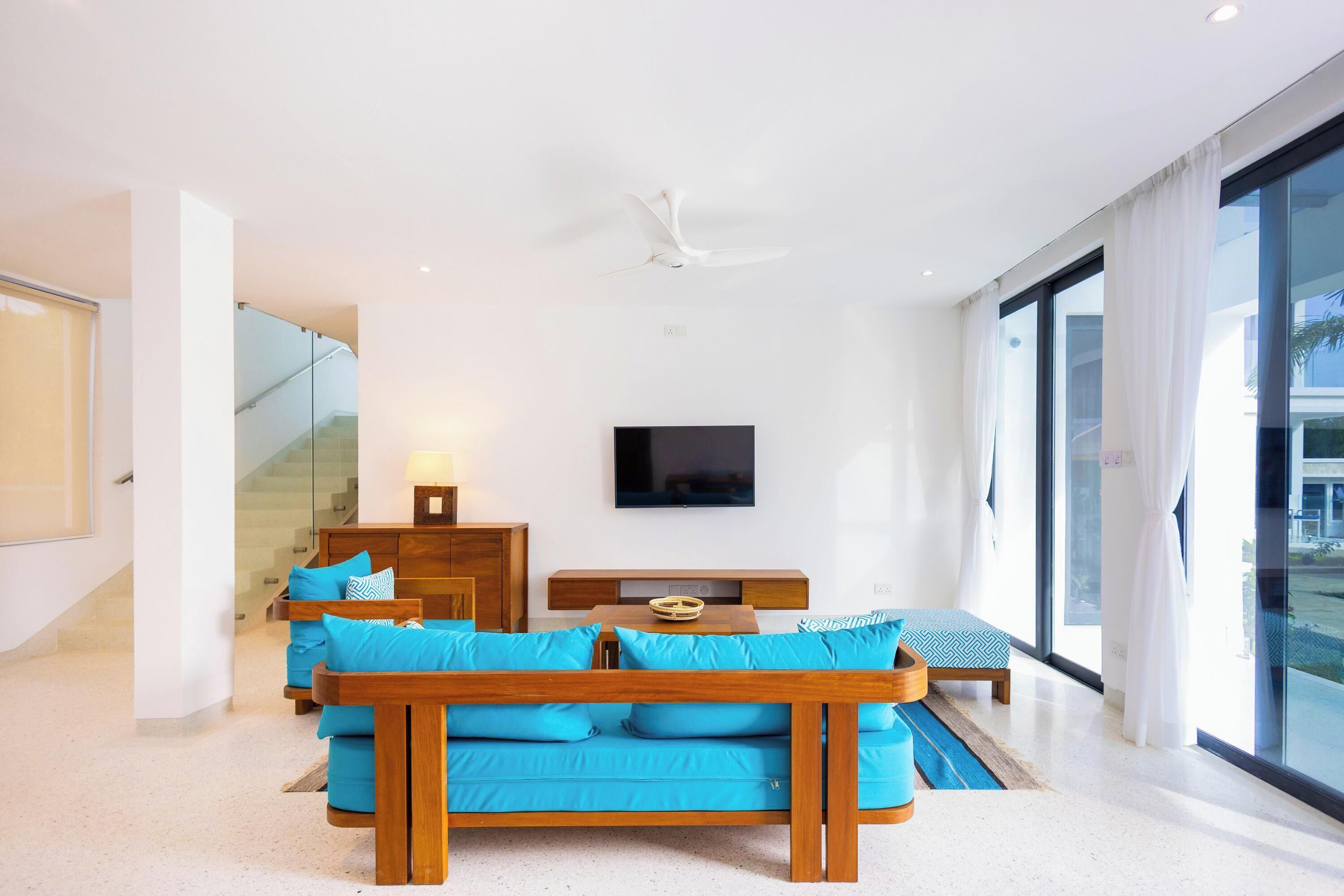 A modern living room with a wooden sofa set, a flat-screen TV, and a glass staircase.
