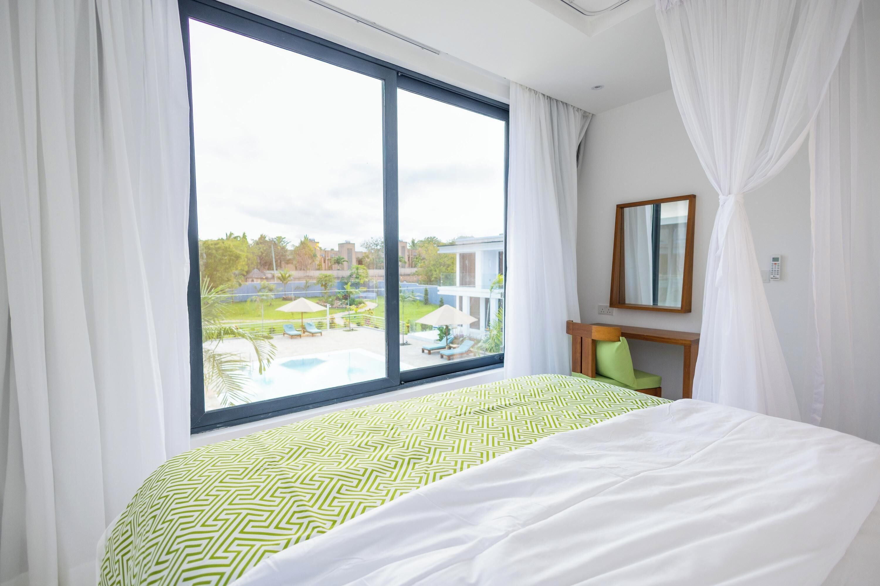 Ett hotellrum med ett stort fönster, en säng med ett mönstrat överkast, en grön stol, ett träbord och en spegel på väggen.