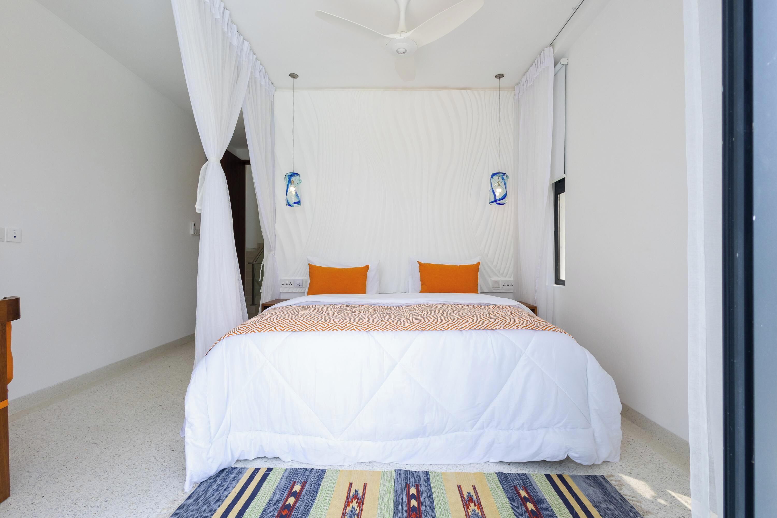 A bedroom with a bed, white curtains, two wall-mounted lamps, and a patterned rug.