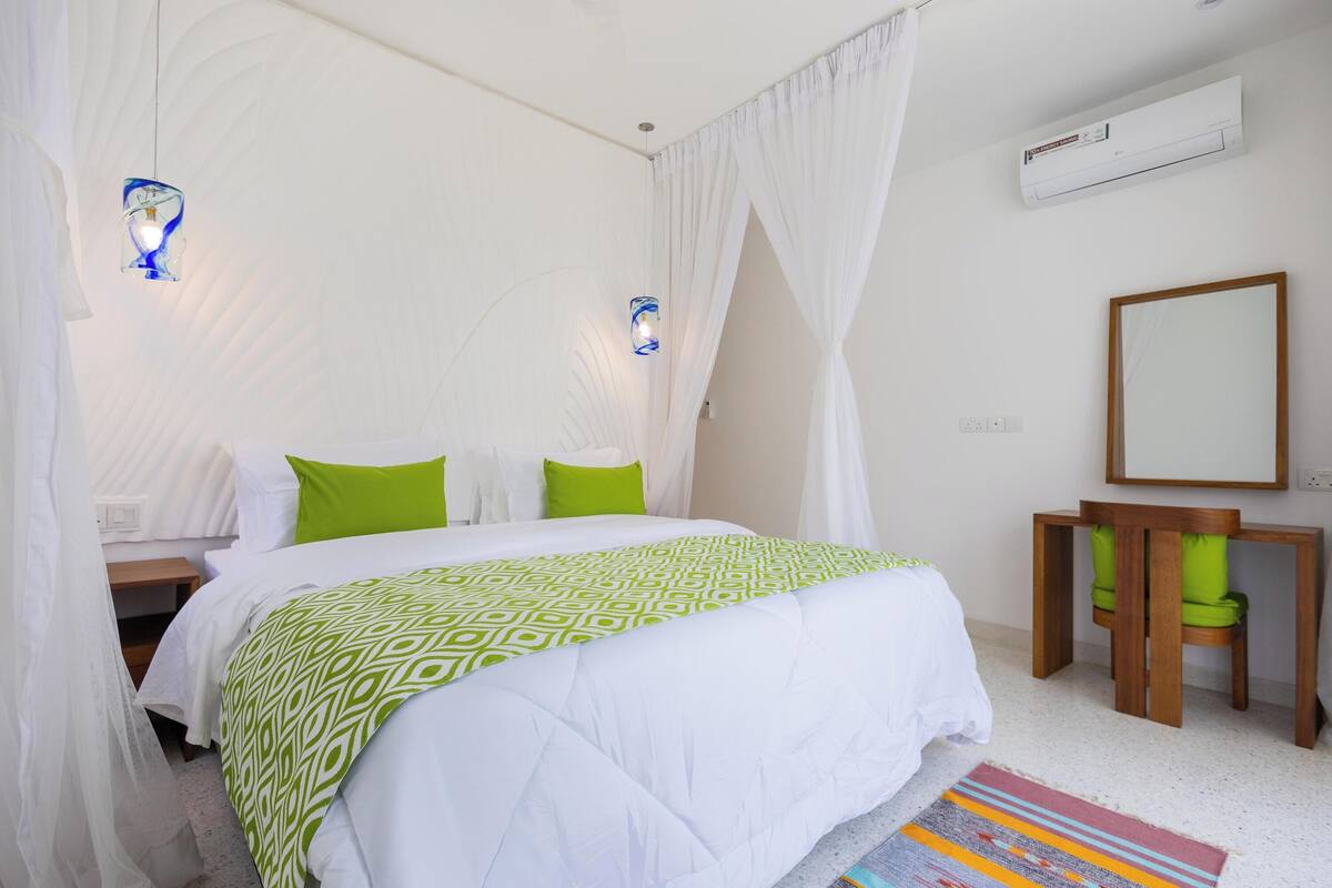 A bedroom with a bed, a bedside table, a chair, a mirror, and an air conditioning unit.