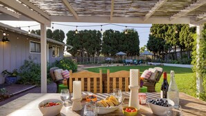 Een terras buiten met een houten tafel gedekt voor een maaltijd, omgeven door groen en een houten hek.