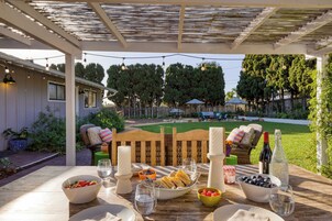 An outdoor patio with a wooden table set for a meal, surrounded by greenery and a wooden fence.