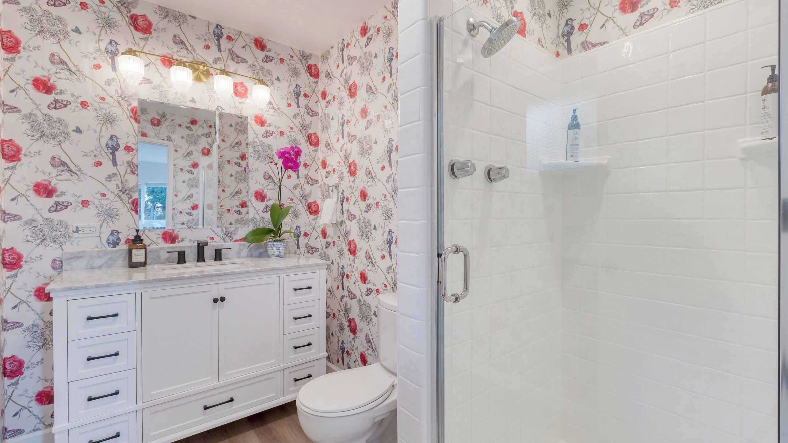 A bathroom with floral wallpaper, a white vanity, a glass shower enclosure, and a white toilet.
