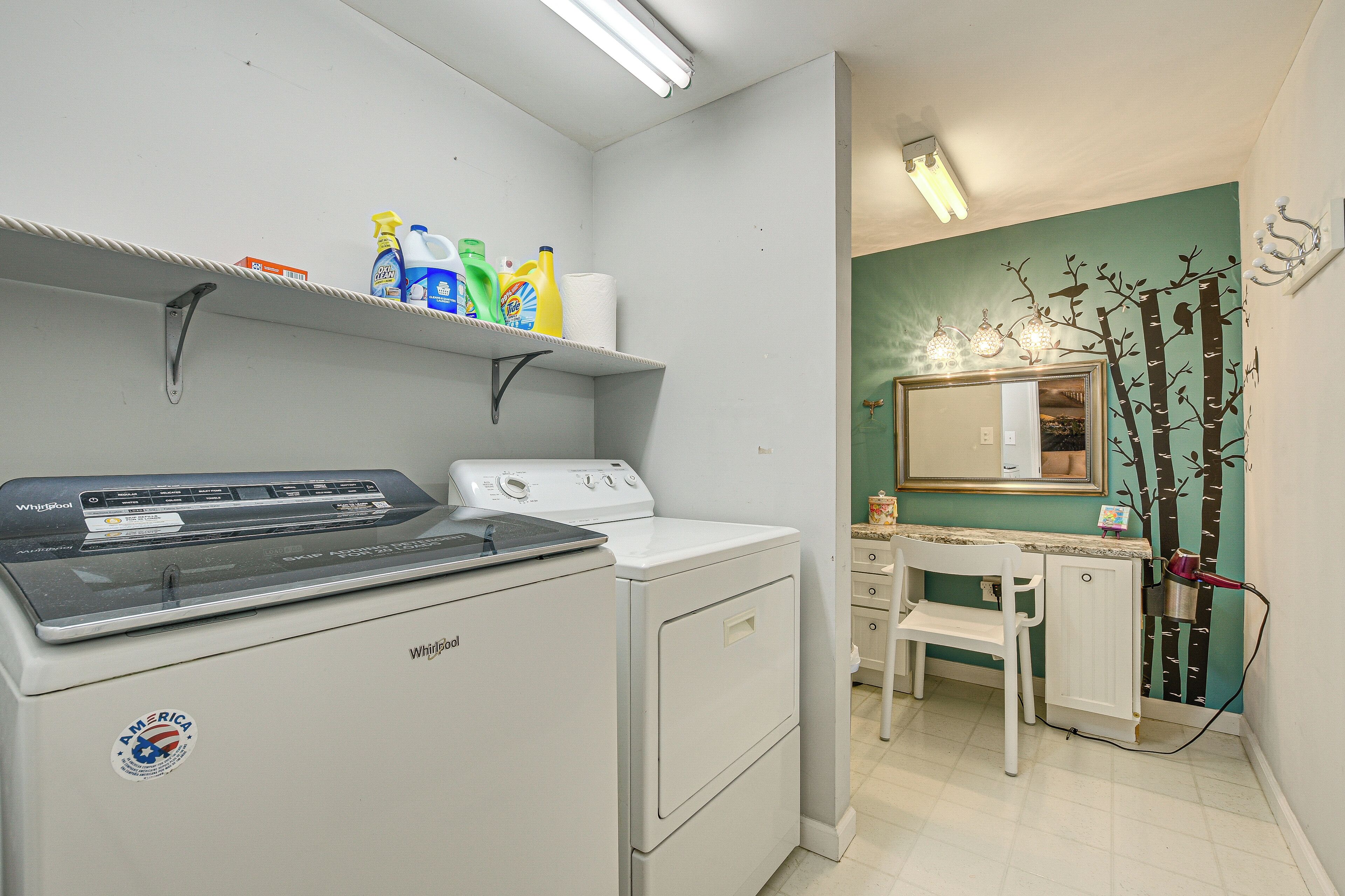 Een wasruimte met een voorlader wasmachine en droger, planken met schoonmaakmiddelen, en een wastafel met spiegel en decoraties aan de muur.