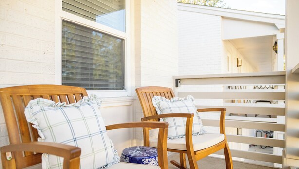 Zwei Schaukelstühle mit karierten Kissen auf einer Veranda mit weißer Reling und einem Fenster mit Rollläden.