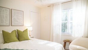 Ein Schlafzimmer mit Bett, grünen Kissen, einem Nachttisch mit Lampe, einem Sessel und einem Fenster mit Vorhängen.