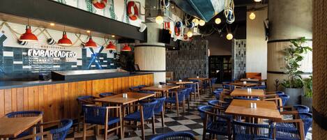 A restaurant interior with wooden tables and blue chairs, a bar area, and a wall decorated with fish and maritime elements.