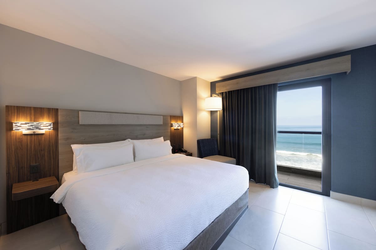 Ett modernt hotellrum med en stor säng, ett nattduksbord, en lampa och utsikt över havet.