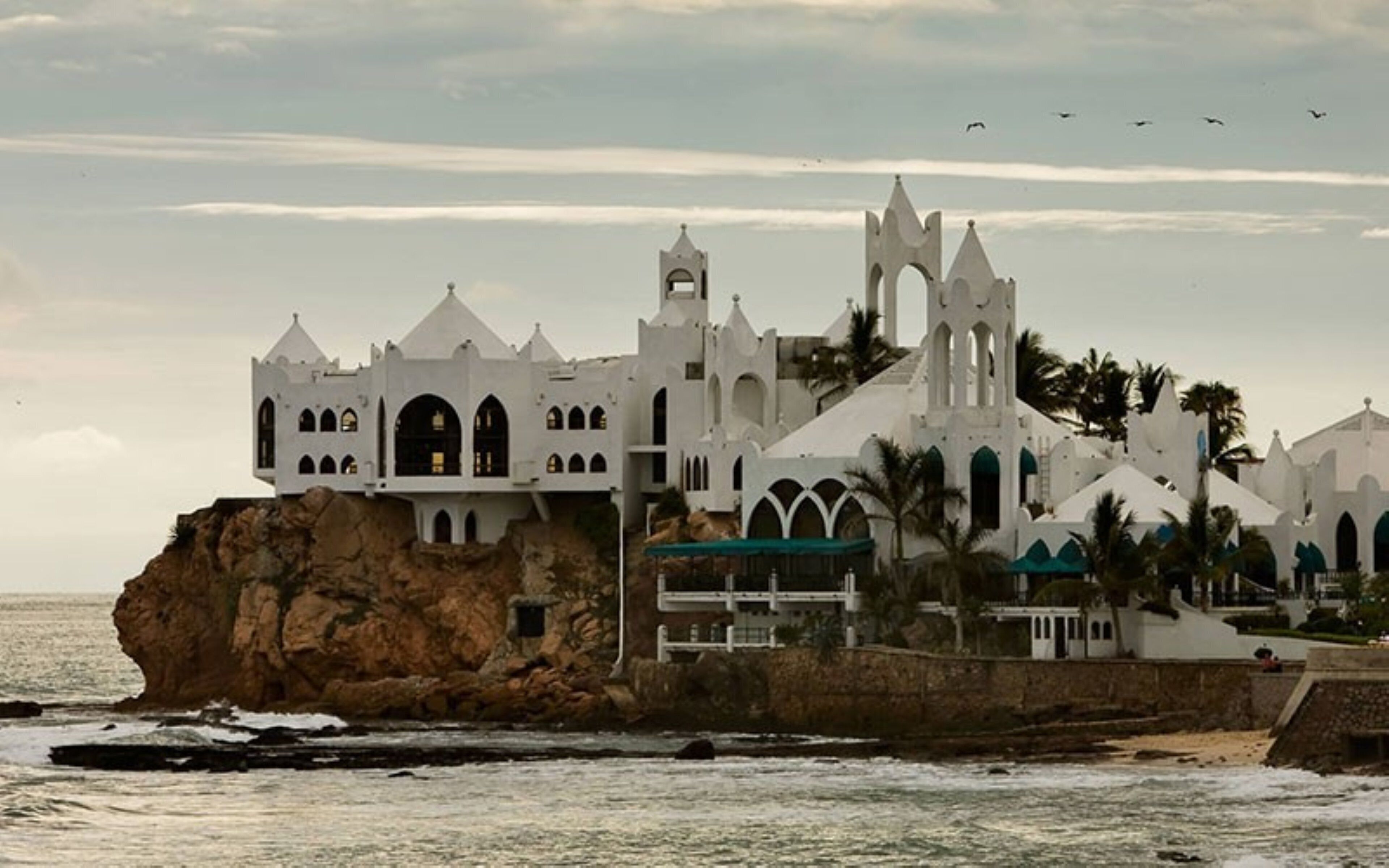 A white building with multiple towers and arches, situated on a rocky outcrop by the sea.